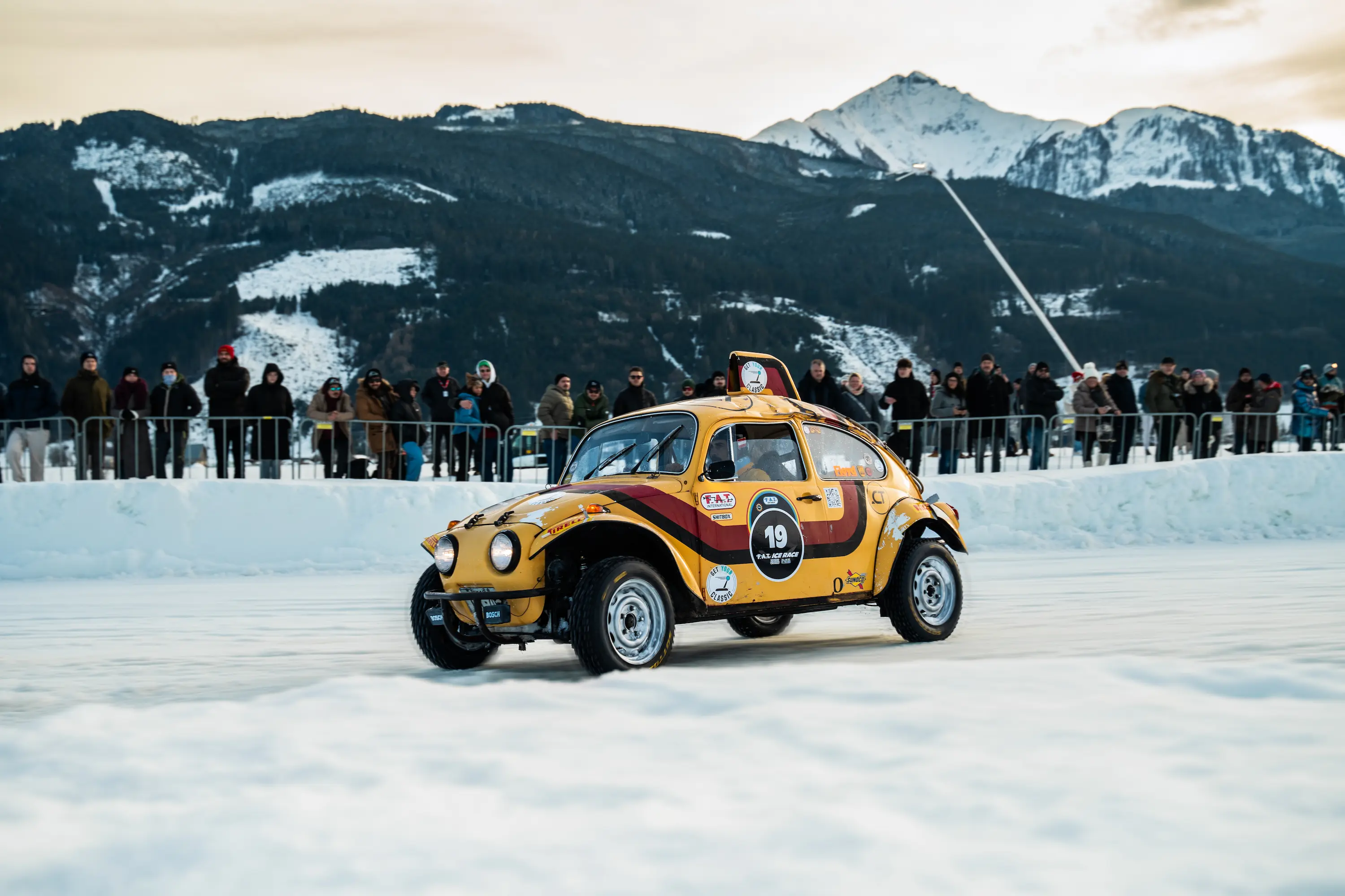 Gold custom-modified VW Beetle number 19 drifting on ice track with spectators and dramatic snow-covered Alpine peaks at FAT Ice Race Zell am See