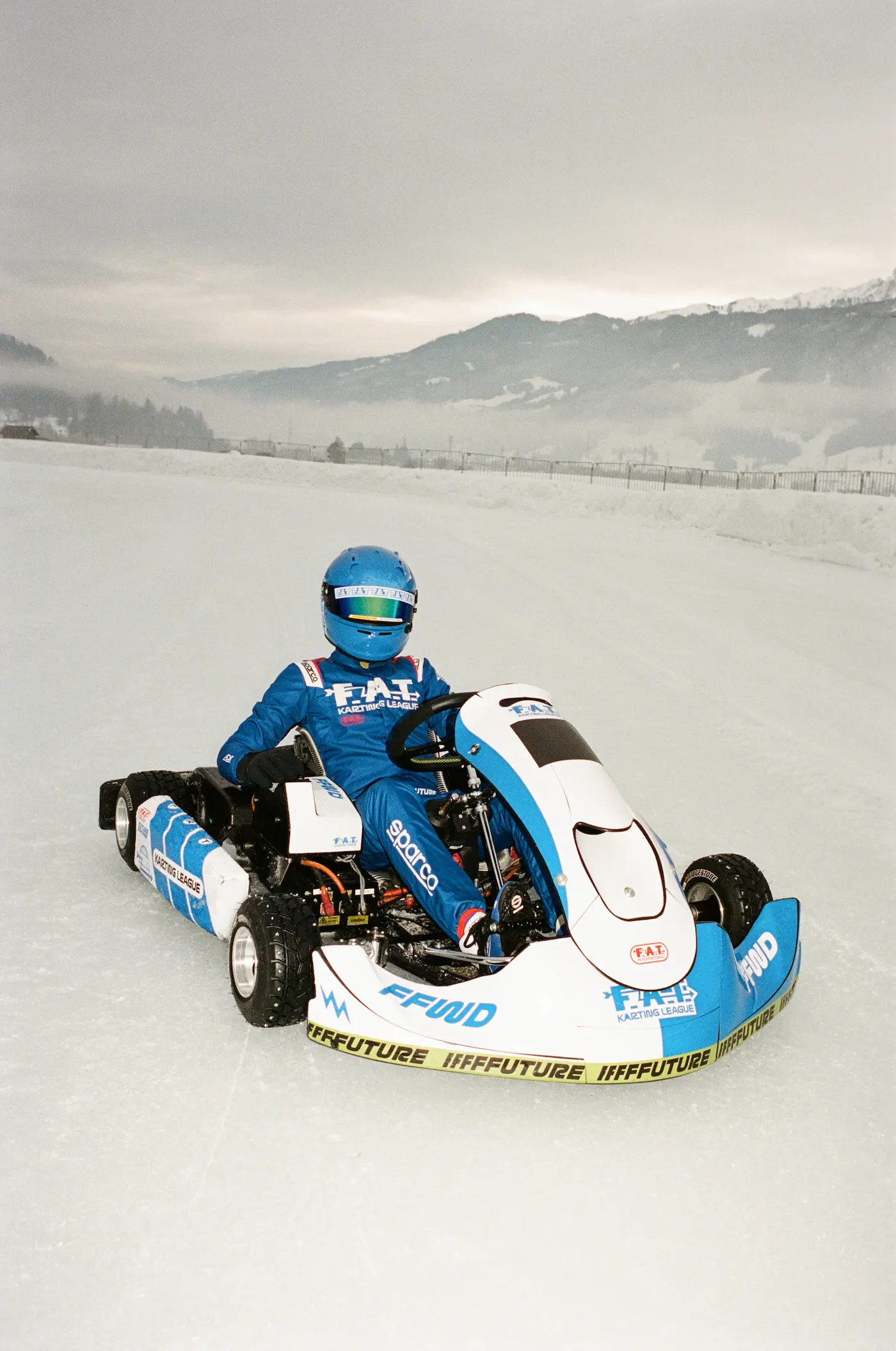 Young driver in blue FAT Racing League suit competing in white ice kart on frozen runway with misty Alpine mountains in background at Zell am See Austria