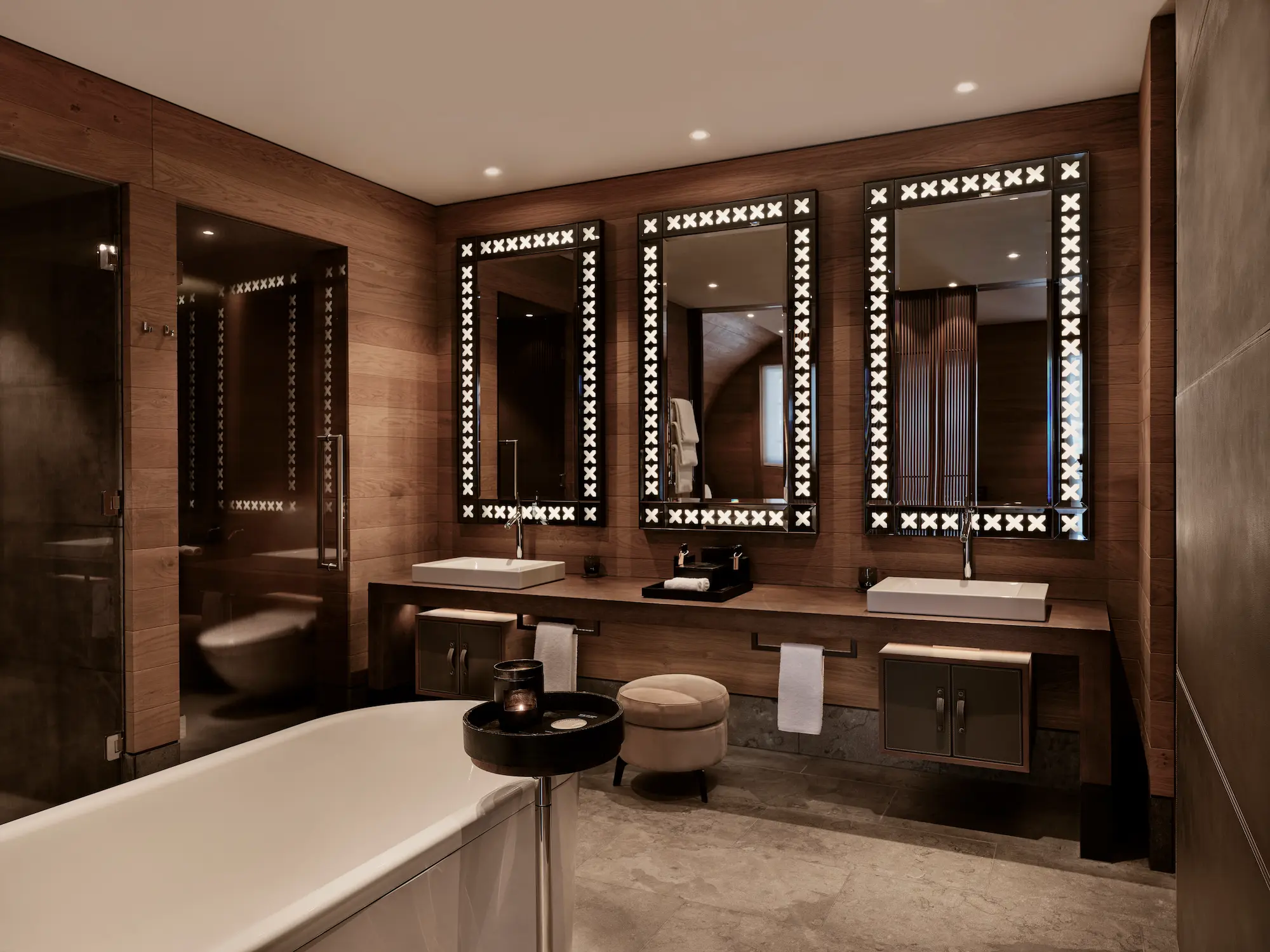 Sophisticated Deluxe Suite bedroom at The Chedi Andermatt with artistic Renaissance-style wallpaper, leather armchair with fur throw, built-in fireplace with timber surround, vertical wood paneling, and luxury bedding by Jean-Michel Gathy