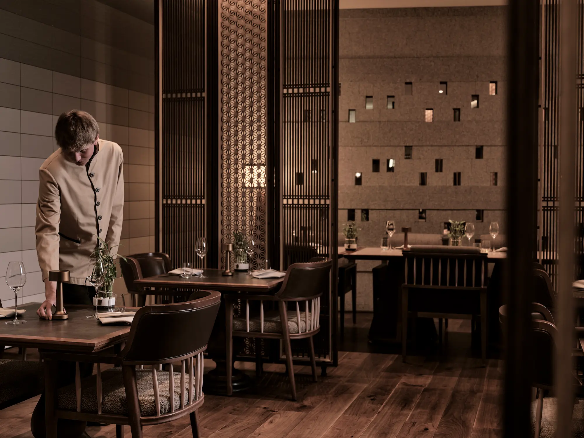 Server in beige uniform setting table at 2-MICHELIN star The Japanese Restaurant, The Chedi Andermatt, featuring intricate geometric wooden screens, textured stone walls with ambient lighting cutouts, wooden dining tables, and refined Japanese minimalist d