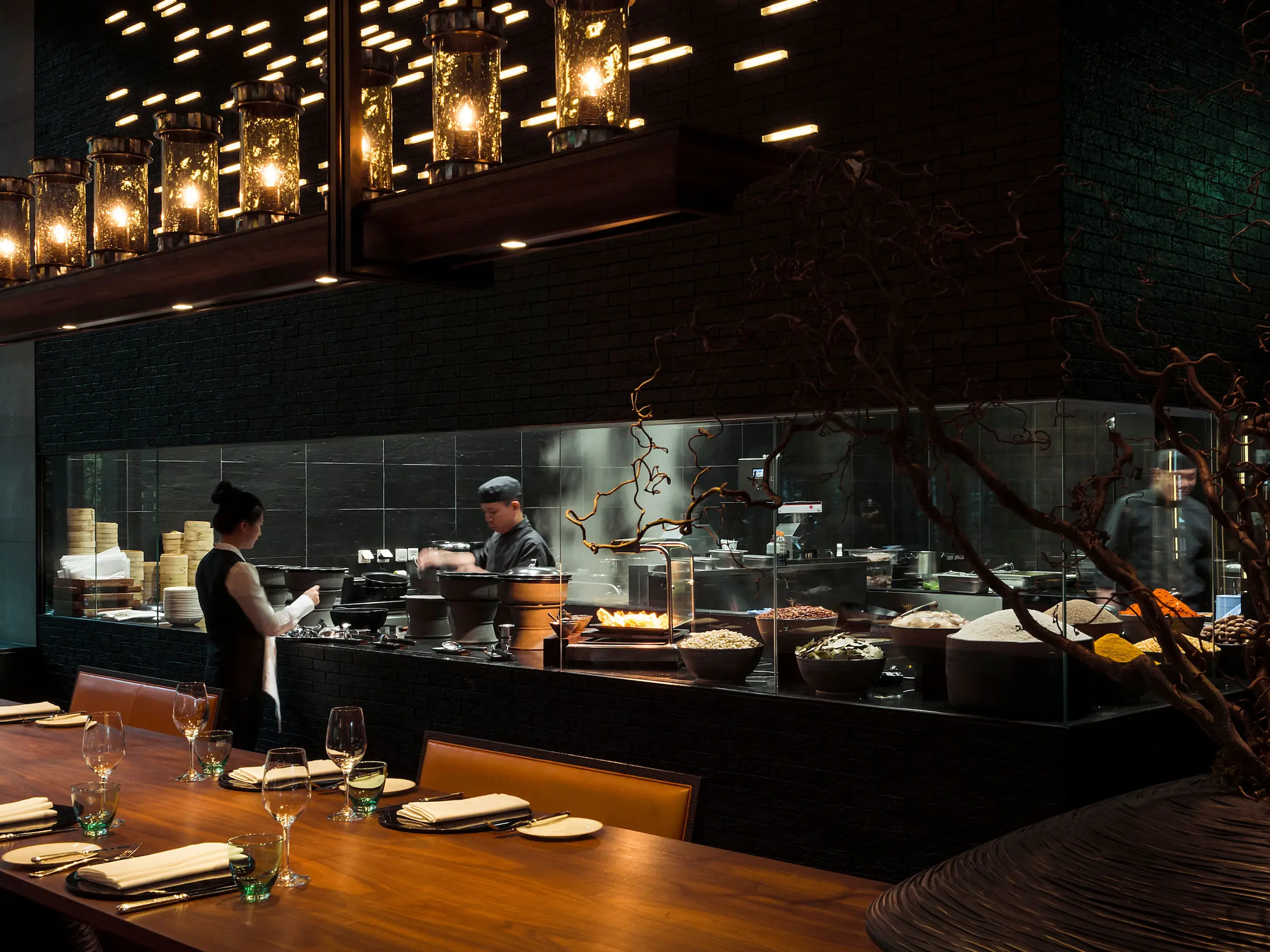 The Restaurant at The Chedi Andermatt featuring dramatic open theatre kitchen with chefs at work, suspended glass lanterns, decorative branch installation, polished wood dining tables, and contemporary Asian-Alpine design by Jean-Michel Gathy