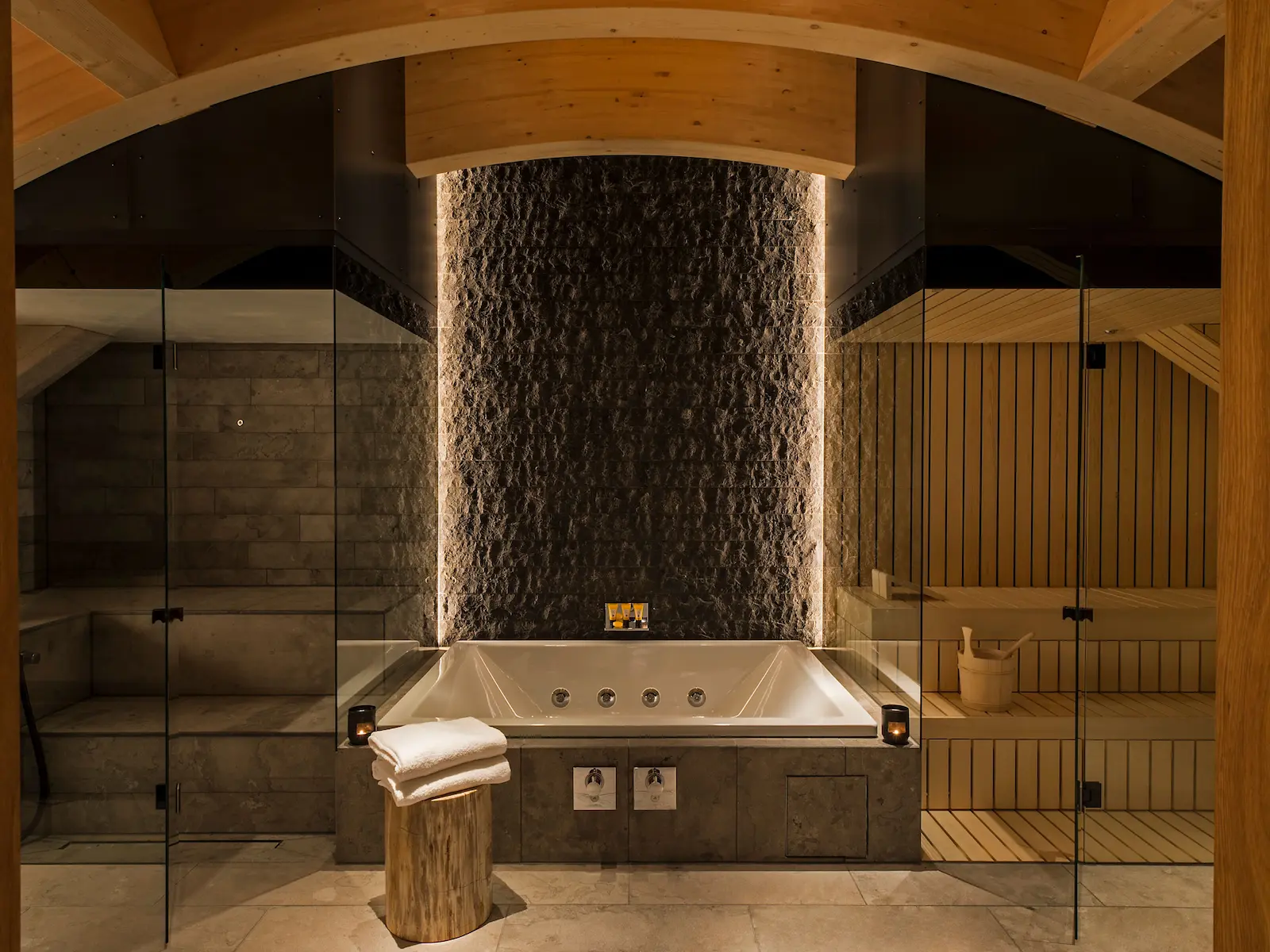 Luxurious Furka Suite private spa at The Chedi Andermatt featuring built-in jacuzzi tub with jet nozzles, backlit textured stone wall, private Finnish sauna with wooden benches, vaulted timber ceiling, glass shower enclosure, and rustic wood stump pedestal