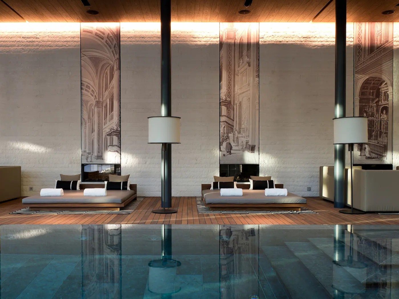 Elegant 35-meter indoor swimming pool at The Chedi Andermatt Spa and Health Club with teak wood deck loungers, sculptural columns, backlit vintage architectural murals, textured stone walls, warm ambient lighting, and serene wellness atmosphere