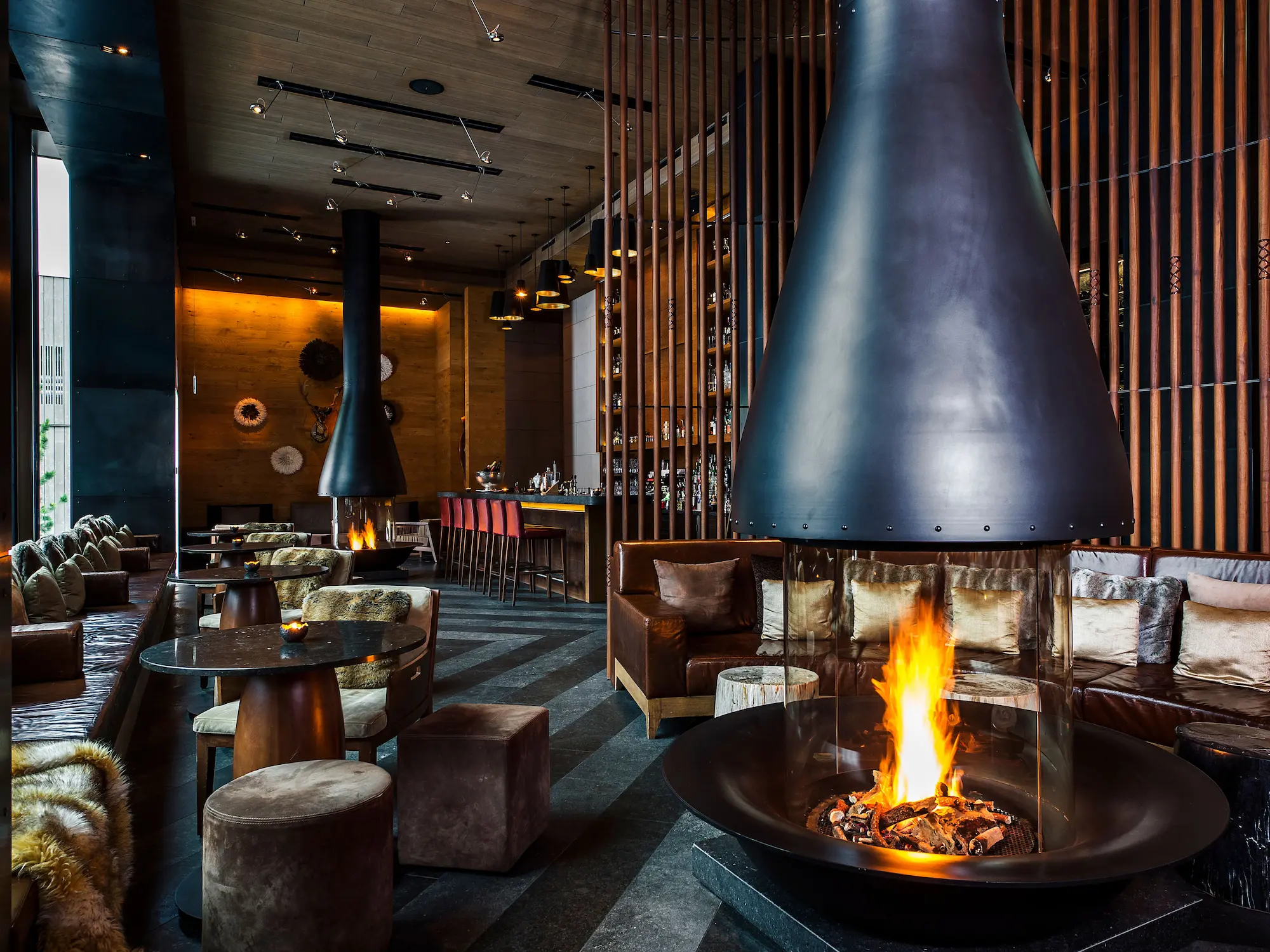 Sophisticated Bar and Living Room at The Chedi Andermatt featuring dual suspended conical fireplaces, leather seating, vertical timber screens, warm ambient lighting, fur throws, and contemporary Asian-Alpine interior design by Jean-Michel Gathy