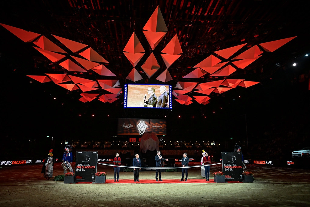 Elegant opening ceremony at Longines CHI Classics Basel 2026 featuring dramatic red geometric LED ceiling installation, officials on red carpet with Longines branding, large video screen, sophisticated arena lighting at St. Jakobshalle