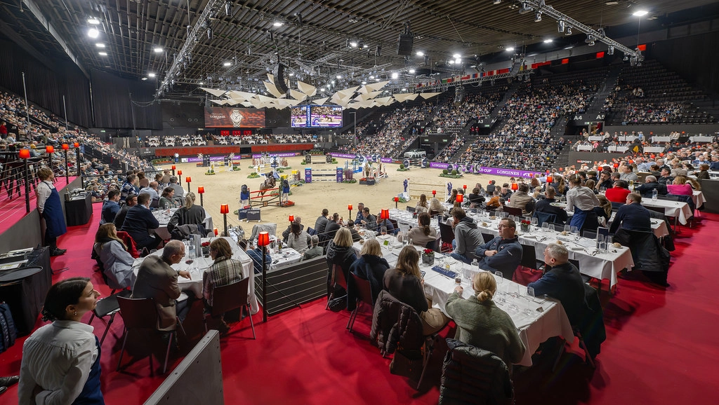 Luxury VIP hospitality seating at Longines CHI Classics Basel with white-tablecloth dining, red carpet, arena-side premium viewing of show jumping competition, packed grandstands at St. Jakobshalle with 25,500 spectators