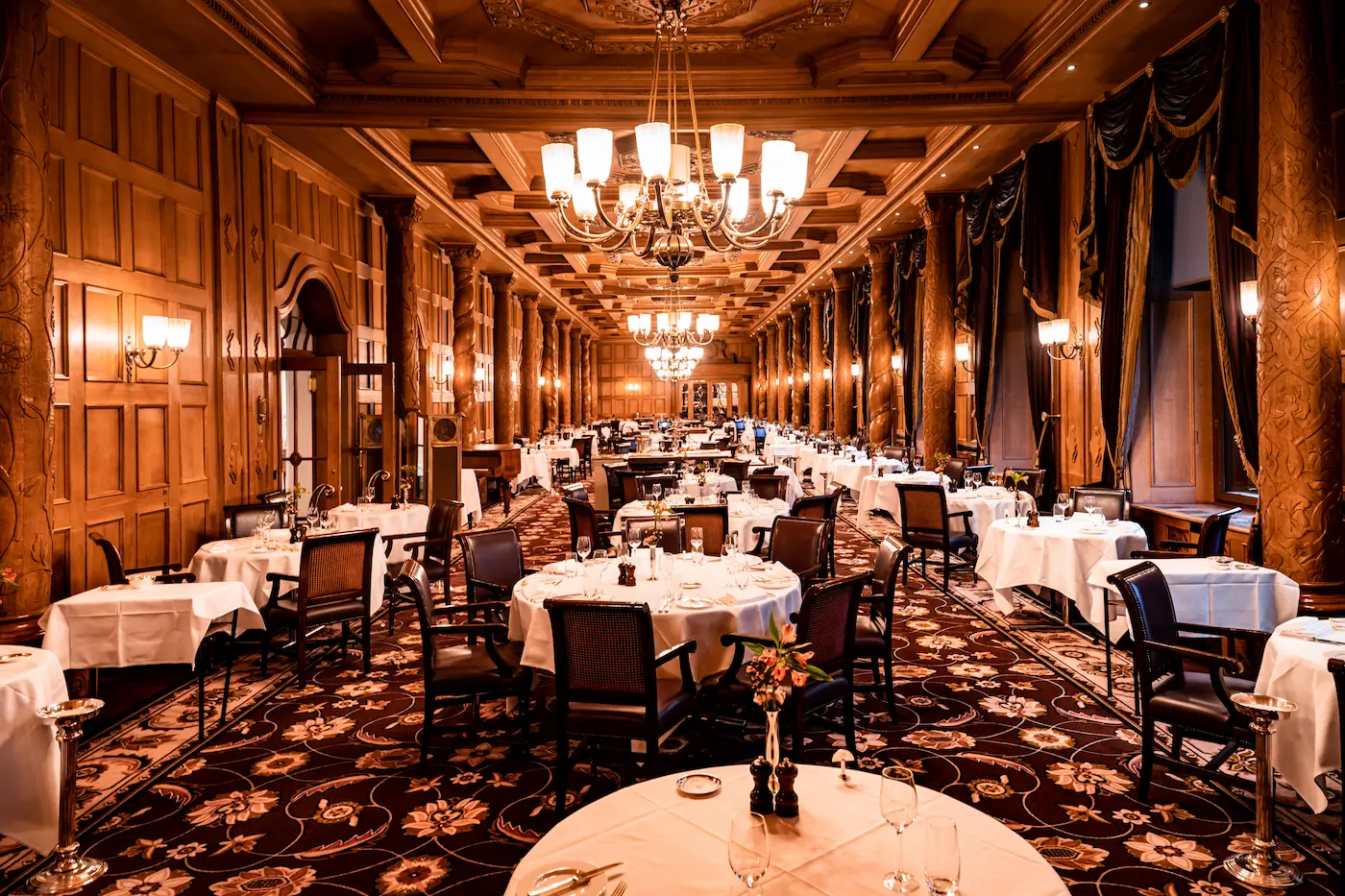 Elegant table setting in Suvretta House Grand Restaurant featuring white linens, crystal glassware, ornate wood paneling, and window view of snow-covered Alpine peaks, showcasing luxury five-star fine dining experience in St. Moritz