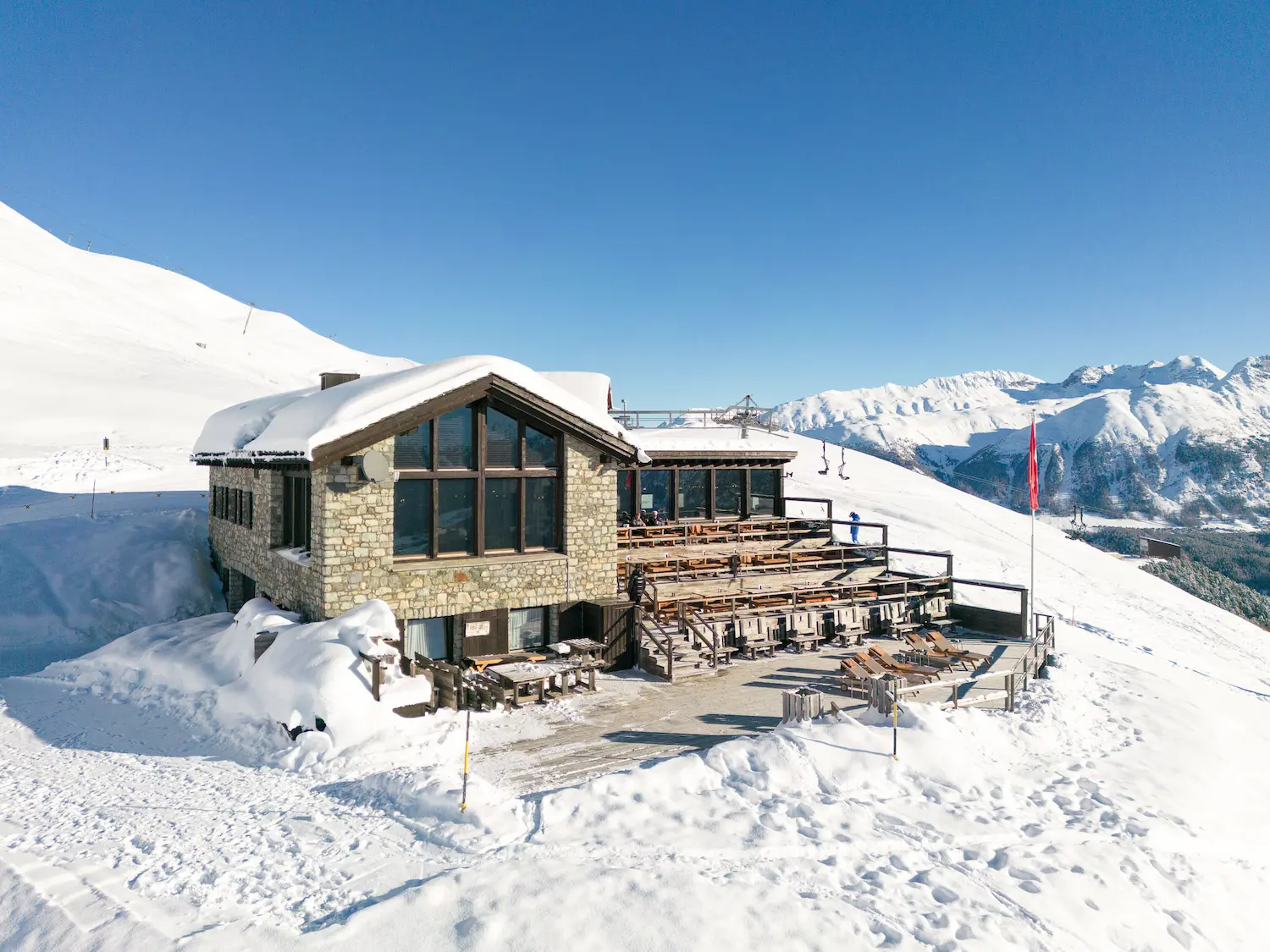 Authentic mountain restaurant chalet at Suvretta House ski area with stone architecture, panoramic terrace seating, deep snow surroundings, and dramatic Alpine peaks backdrop, offering exclusive slope-side dining experience in St. Moritz