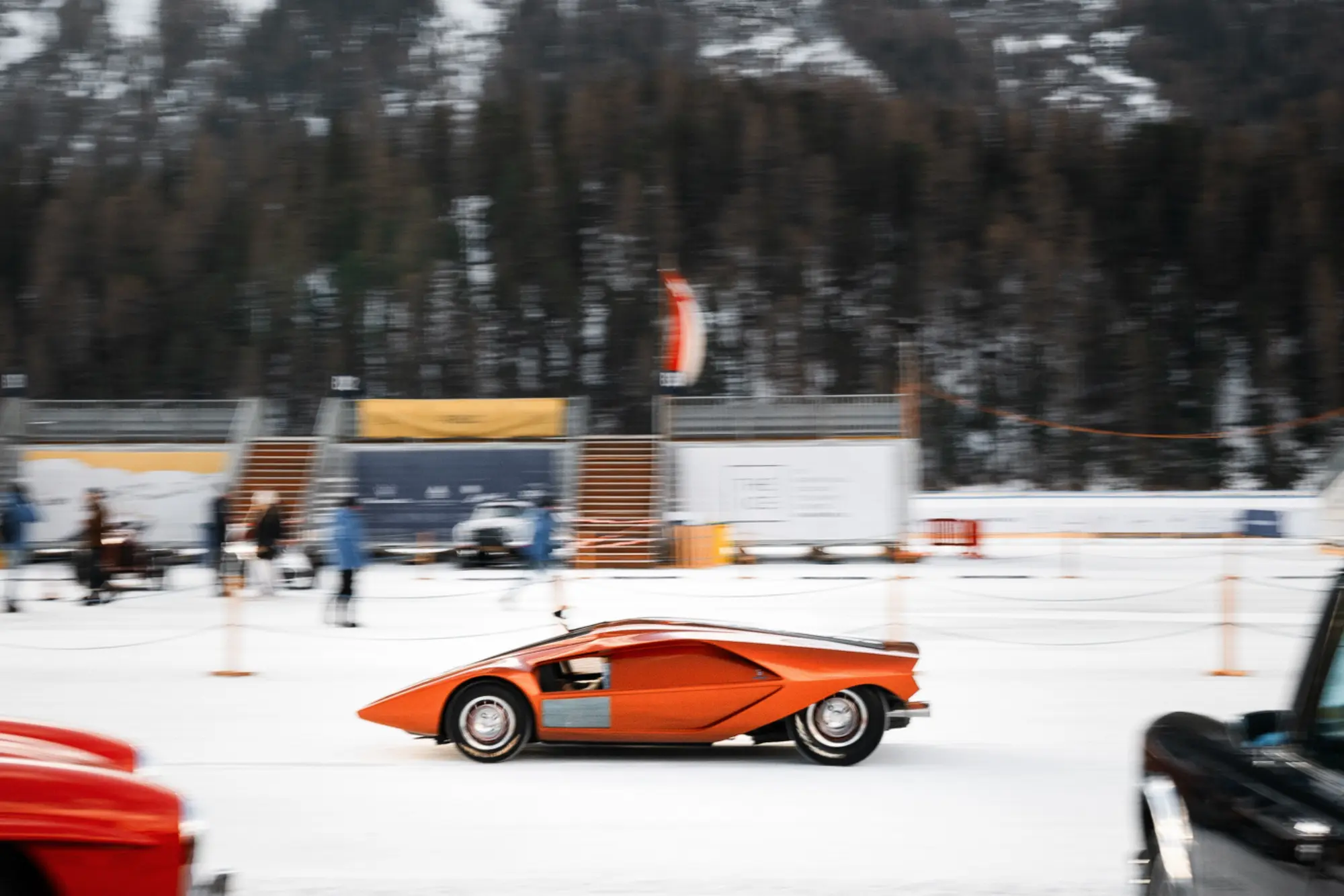 Orange Lancia Stratos Zero concept car in motion on frozen Lake St. Moritz at The ICE International Concours of Elegance with dynamic driving display, spectators and sponsor pavilions along ice track, pine forest backdrop showcasing on-ice automotive perfo