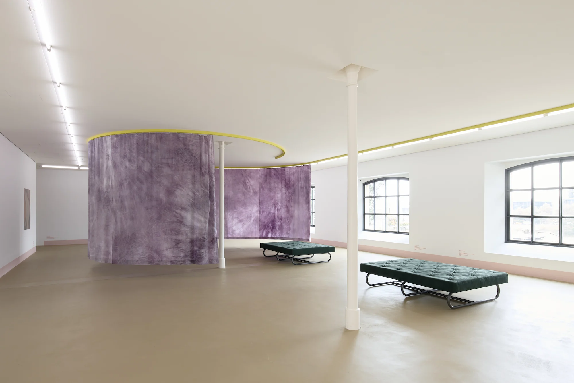 Minimalist contemporary art installation in white museum gallery featuring suspended yellow circular track system with purple translucent curtain divider, green tufted daybed benches, arched windows with natural light, and beige resin flooring showcasing f