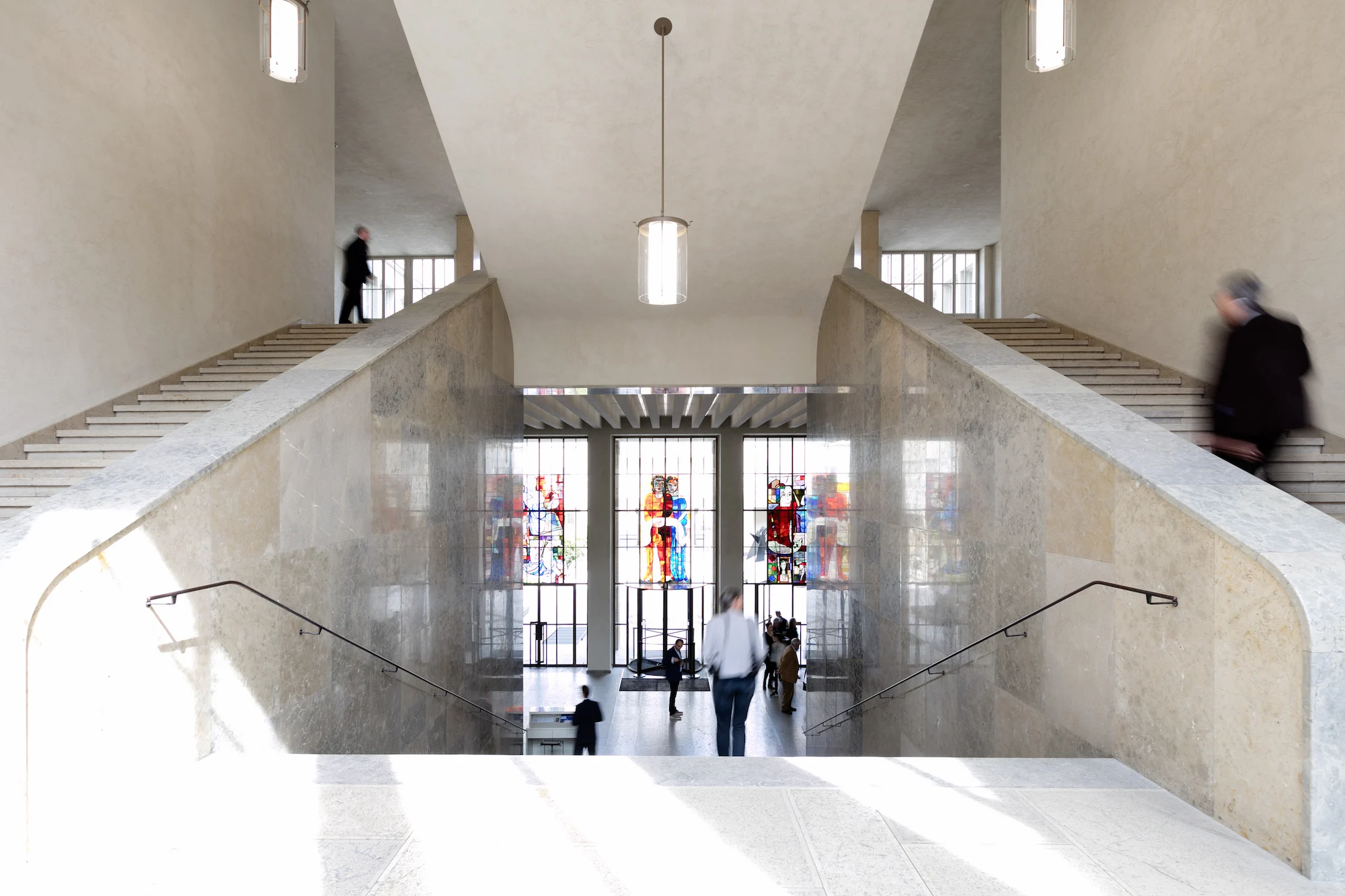 Monumental symmetrical grand staircase at Kunstmuseum Basel with beige limestone walls, dual ascending staircases, coffered ceiling with cylindrical pendant lights, colorful stained glass windows visible at ground level entrance