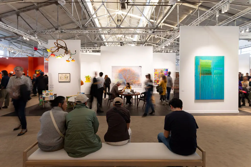 Busy main floor at FOG Design+Art 2026 Fort Mason Center Festival Pavilion showing collectors seated on white bench viewing gallery booths with contemporary artworks including turquoise abstract painting