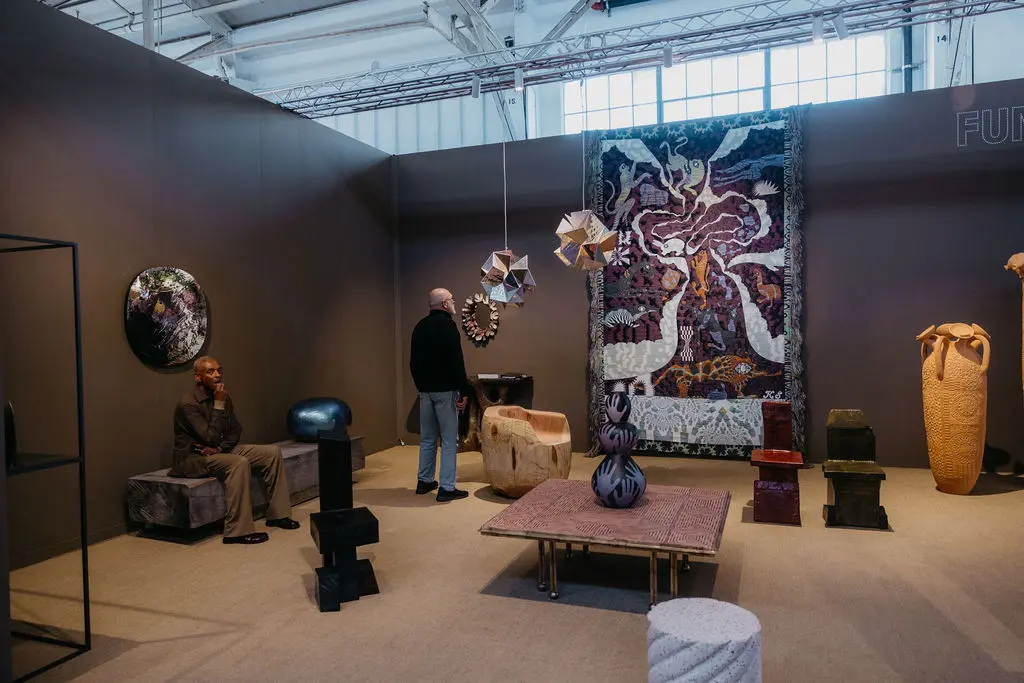 Sophisticated gallery booth at FOG Design+Art 2026 Fort Mason with charcoal gray walls displaying large contemporary textile tapestry with figurative motifs, ceramic vessels, sculptural furniture including carved wood chair and table