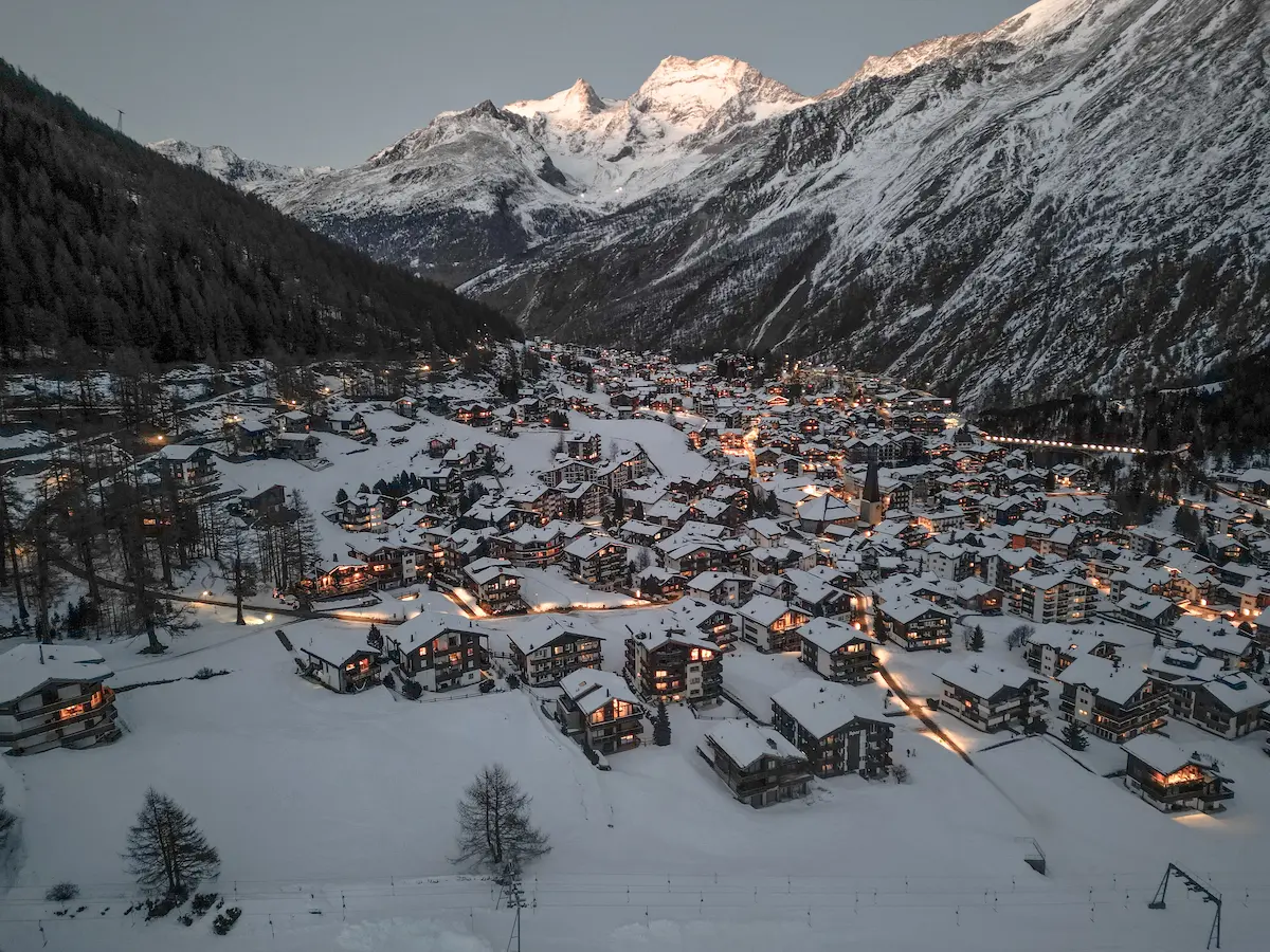 Aerial view of car-free Saas-Fee village at dusk with snow-covered traditional Swiss chalets illuminated by golden lights nestled in Saastal Valley beneath dramatic four-thousanders peaks including Dom