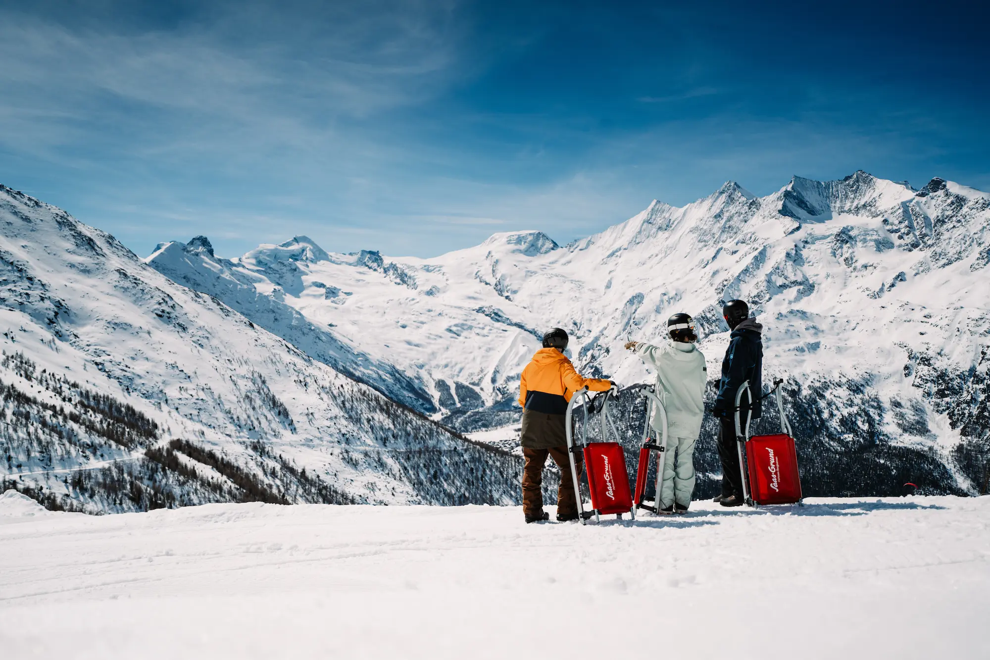 Three people with red sleds standing on pristine snow slope overlooking spectacular panorama of 18 four-thousanders peaks including glaciers in Saas-Fee Saastal Valley Switzerland