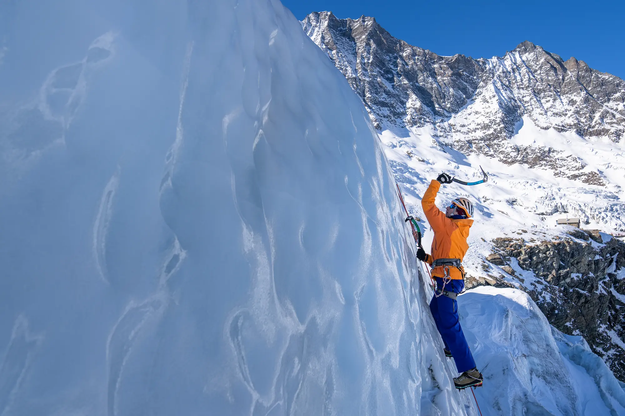 Ice climber in orange jacket ascending vertical blue glacier wall with ice axes and crampons at Saas-Fee with dramatic four-thousanders peaks backdrop