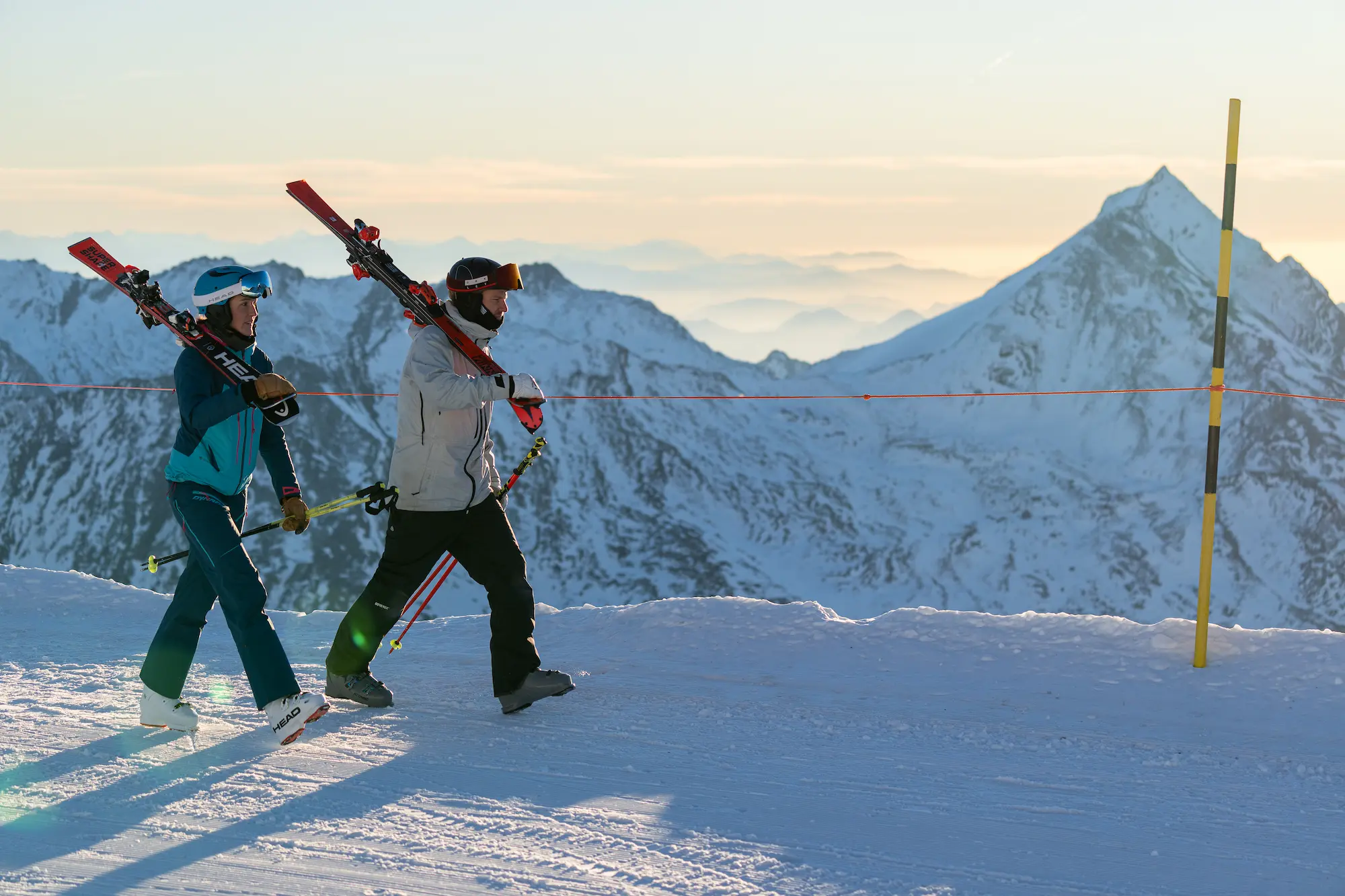 Two skiers with equipment walking on groomed slopes at dawn at Saas-Fee with spectacular panorama of 18 four-thousanders peaks including Dom in pastel sunrise light