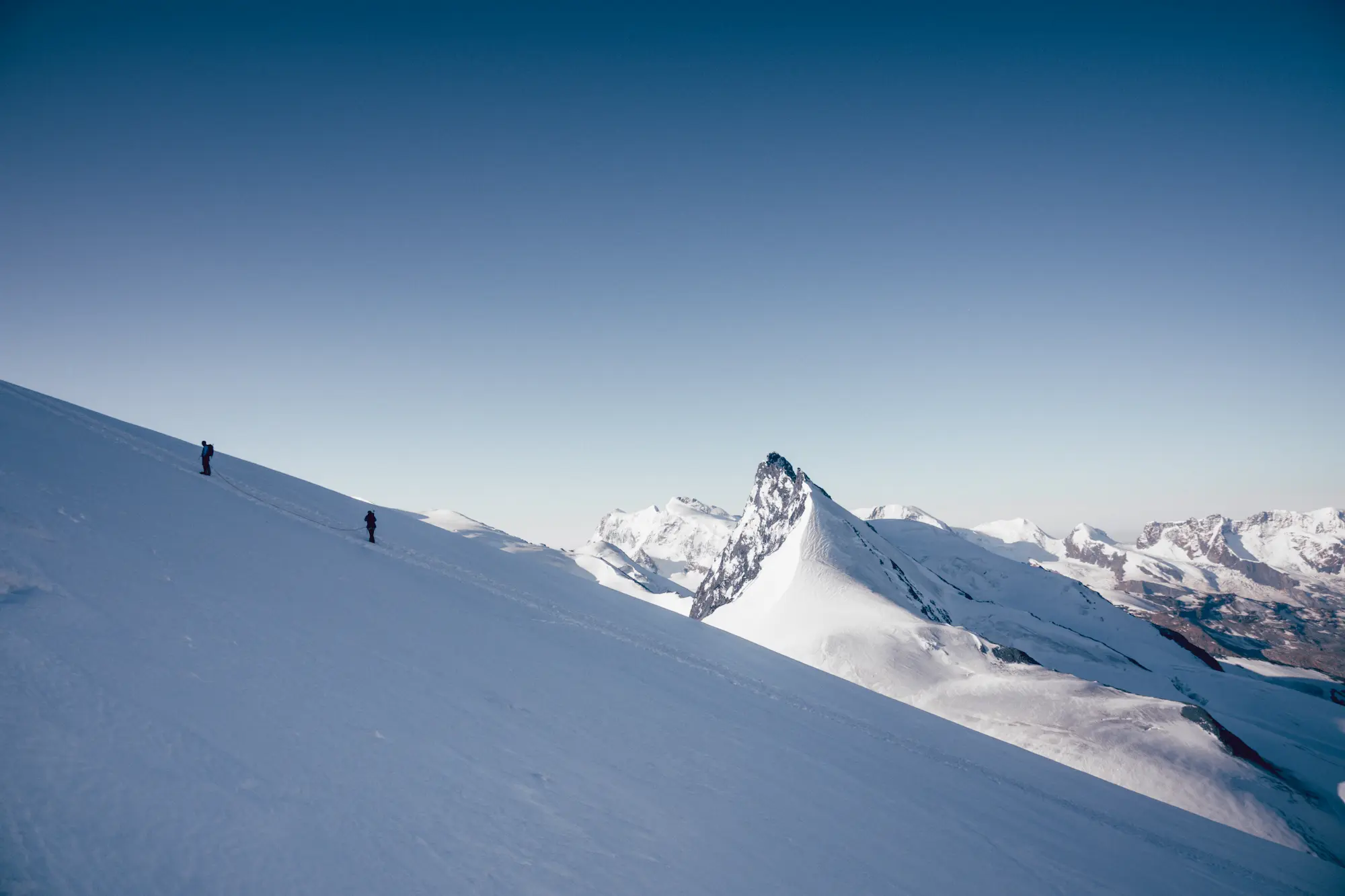 Two roped mountaineers ascending steep snow slope toward Allalinhorn 4,027-meter summit at Saas-Fee with dramatic four-thousanders peaks panorama and clear blue sky