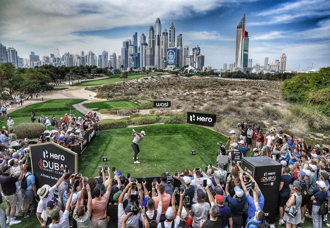 Professional golfer teeing off at the Hero Dubai Desert Classic surrounded by enthusiastic spectators at Emirates Golf Club's signature desert-style Majlis Course hole