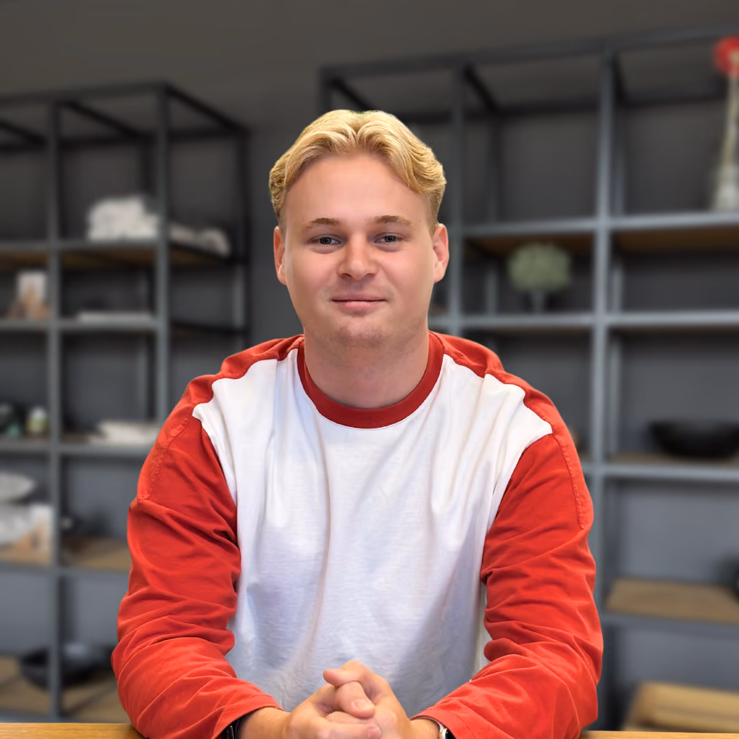 Jonge man met blond haar en rode en witte trui zit met gevouwen handen voor zich, met grijze rekken en decoraties op de achtergrond.