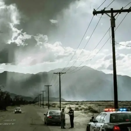Police officer conducting a roadside sobriety test with a man beside a car on a desert road near Indio, California with mountains and telephone poles in the background.