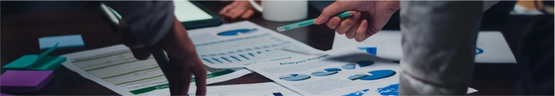 Two people analyzing printed charts and graphs on a desk, one pointing with a pencil.