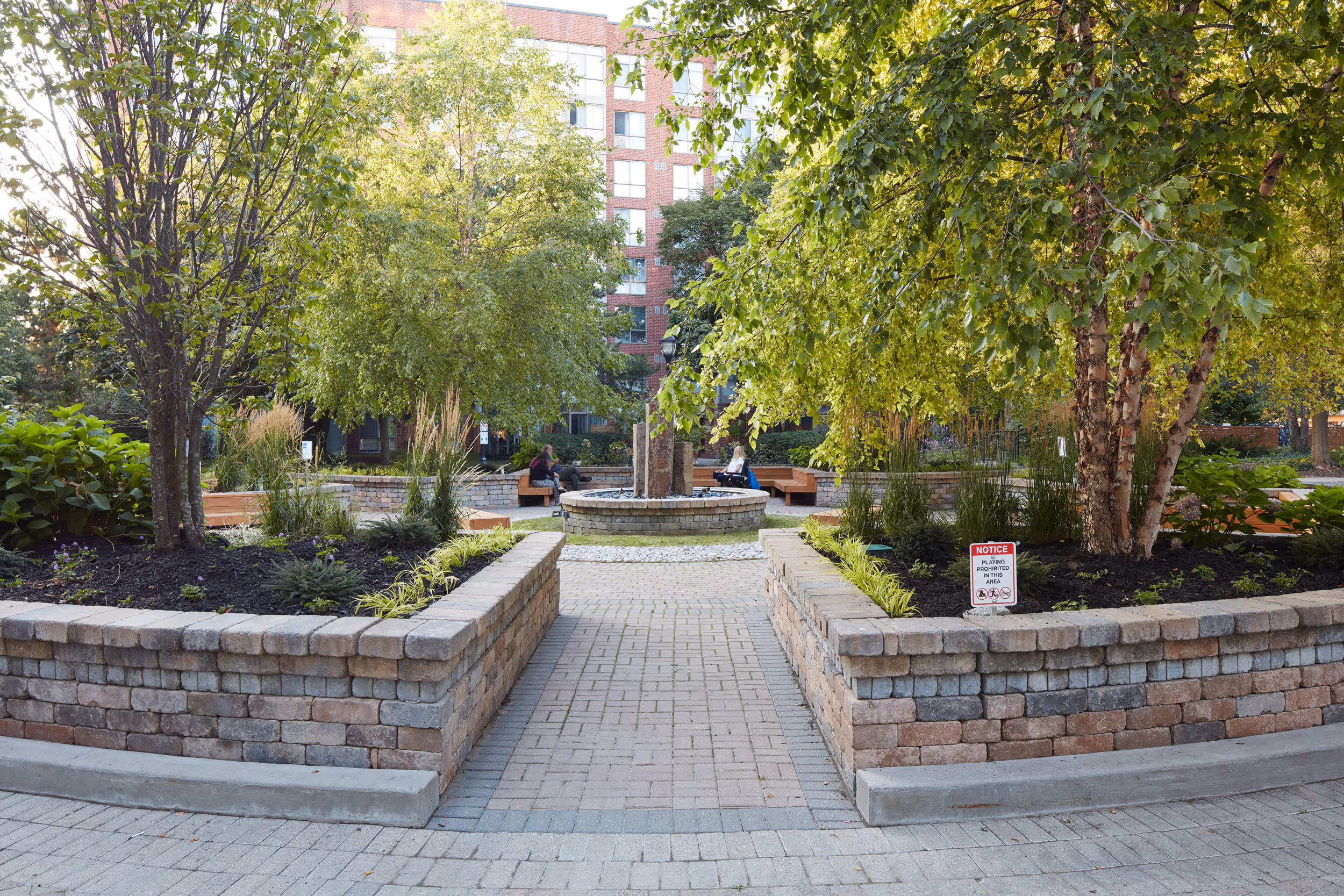 Stone Retaining walls flank entranceway to garden communal area. Garden beds contain trees and perennials. 