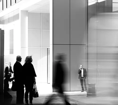 Black and white photo of people standing and walking in a modern urban area, with one man speaking on a phone near a wall.