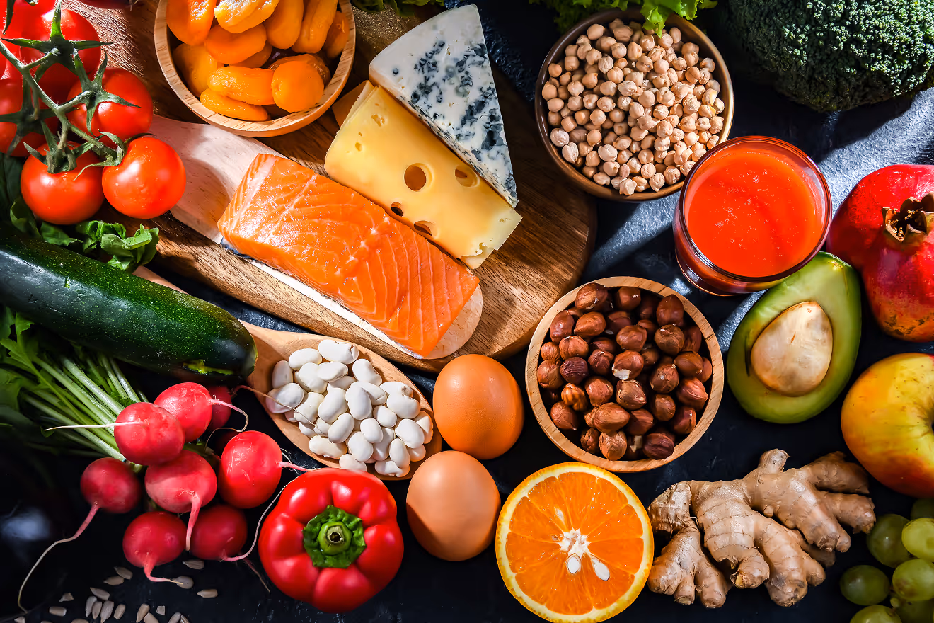 An assortment of healthy foods including salmon, different types of cheese, cherry tomatoes, dried apricots, chickpeas, radishes, red bell pepper, beans, eggs, orange half, avocado, ginger, broccoli, apple, and grapes.