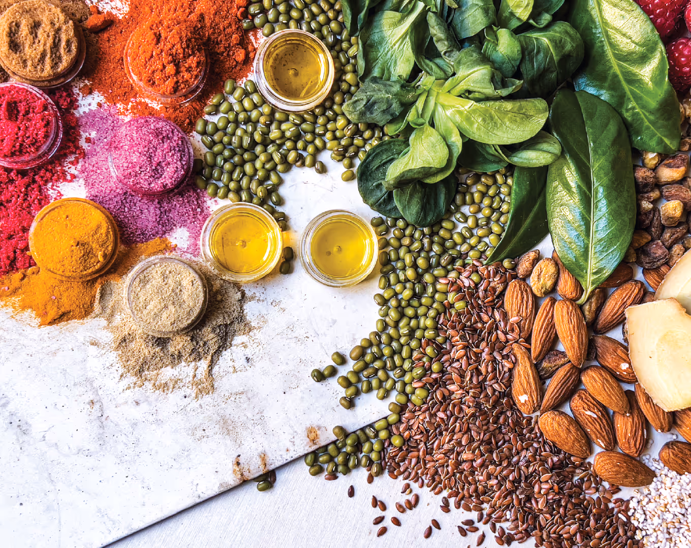Assorted spices in small glass bowls, green mung beans, flax seeds, almonds, leafy greens, slices of ginger, and small cups of oil arranged on a white surface.