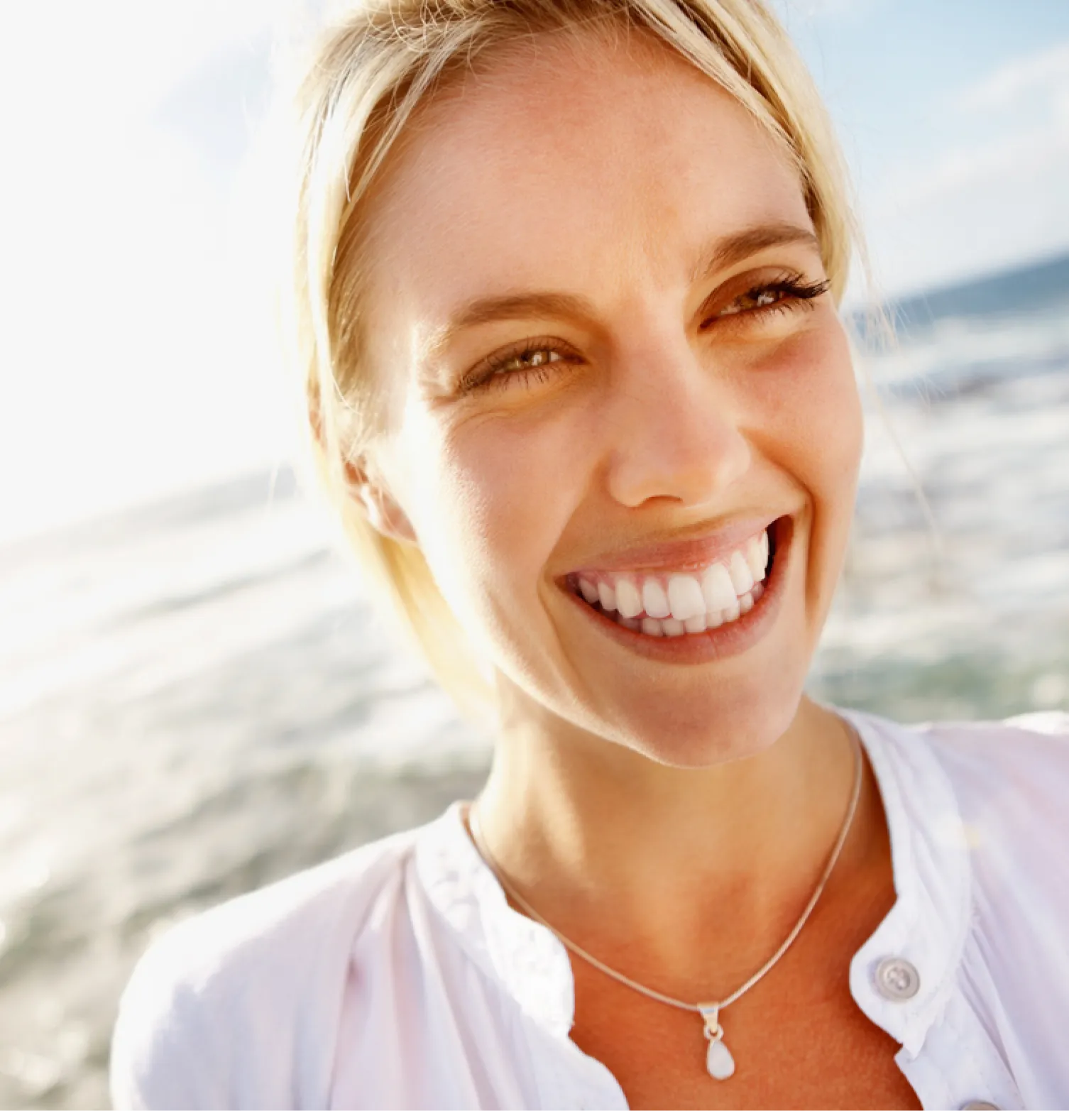 A woman with blonde hair is smiling widely near a beach.