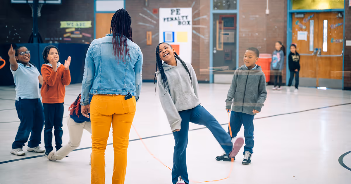 An Upper Academy teacher is interacting with their scholars in the gymnasium. The Upper Academy student in focus is standing on one foot and smiling at her.
