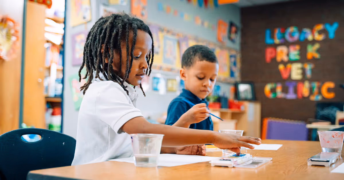 Two Primary Academy students are painting at a shared table. Brightly colored letters spell out "Legacy Pre K Vet Clinic" in the background.