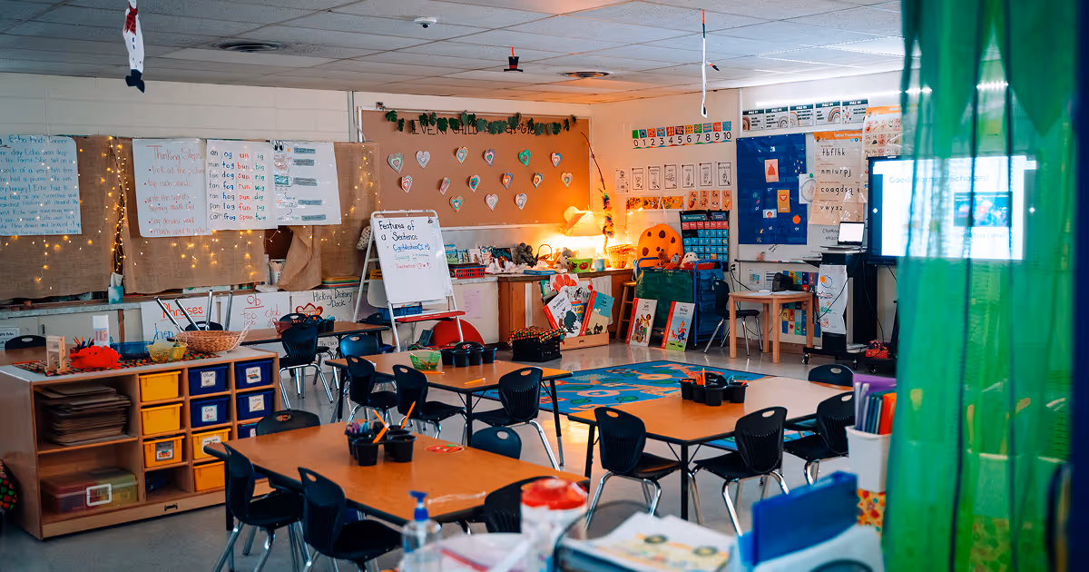 An empty, open classroom with string lights and warm lighting. Several shared desks are doted around the room with a reading rug in the corner.