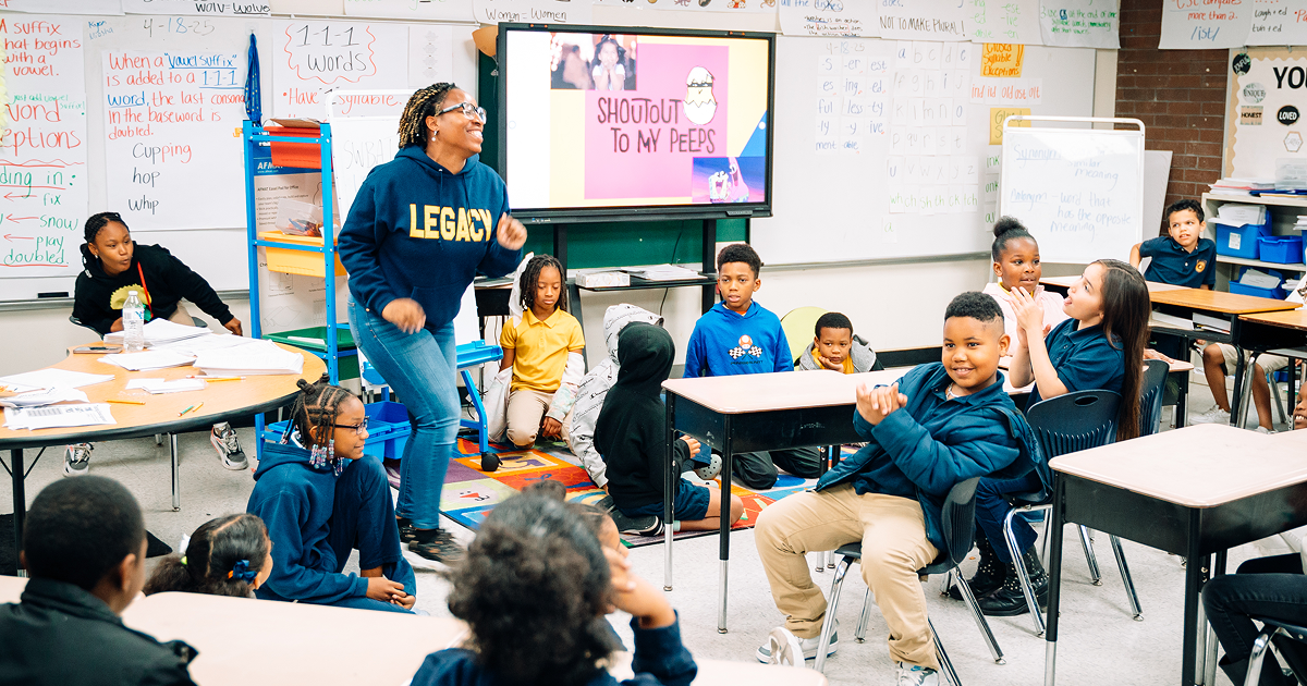 A TLCS teacher wearing a navy "LEGACY" hoodie is dancing in front of a class of Primary Academy scholars. 