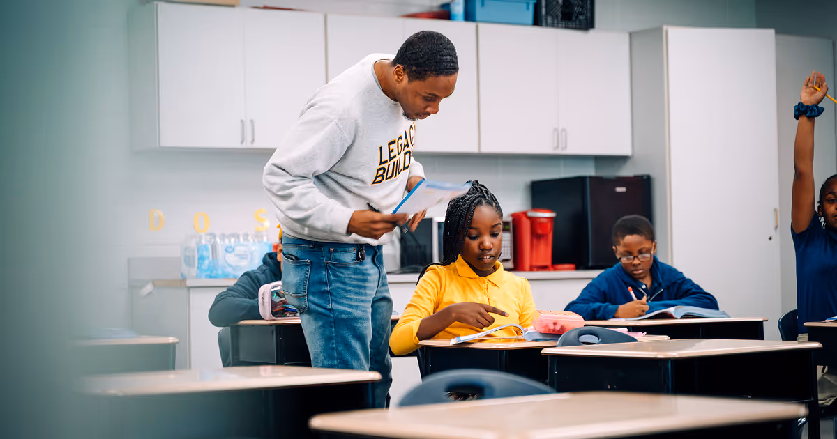 A TLCS teacher wearing a gray "Legacy Builder" sweatshirt is looking over the shoulder of an Upper Academy scholar pointing at her book.