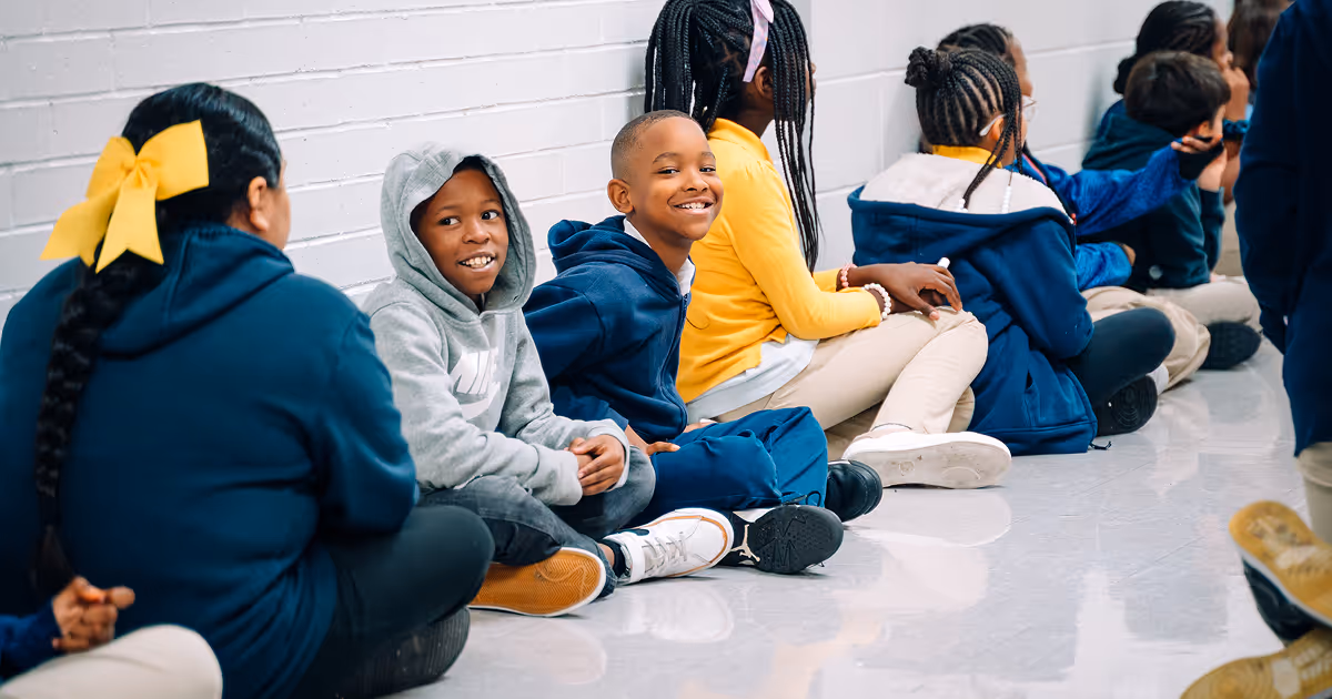 A row of Upper Academy scholars are sitting along a hallway wall. Two scholars are grinning at the camera.