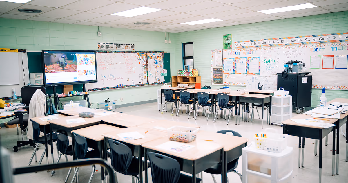 An empty TLCS classroom with clusters of desks around the room with empty floor space in the center. A TV is at the front of the class. 