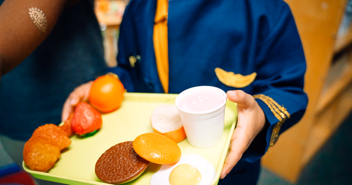 A Primary Academy scholar is holding a toy tray of plastic "Play Food" towards the camera. The tray includes a toy hamburger, strawberry, orange, egg, drink, bread, and chicken nugget.