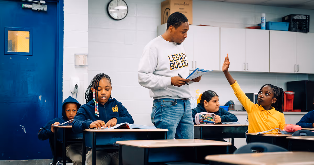A Tulsa Legacy scholar has her hand up to ask a question to the teacher who is walking around. He is wearing a gray "Legacy Builder" sweatshirt on.