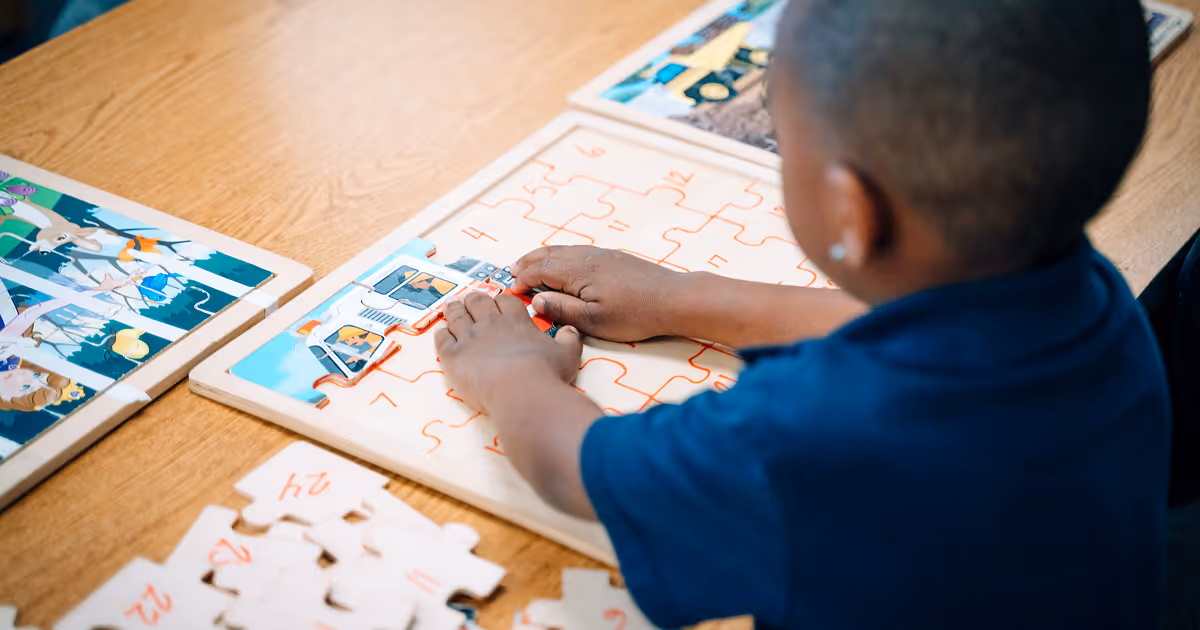 A Primary Academy scholar is arranging wooden puzzle pieces in a puzzle board on a table.