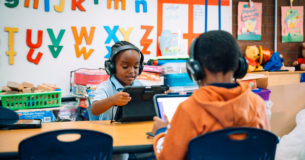 Two preschool scholars wearing headphones and playing on iPads. The background has brightly colored letters of the alphabet.