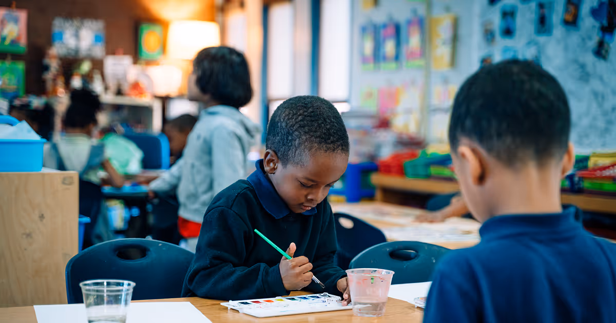A Pre-K scholar is using a green paintbrush to paint with watercolors. Other scholars are standing in the background.