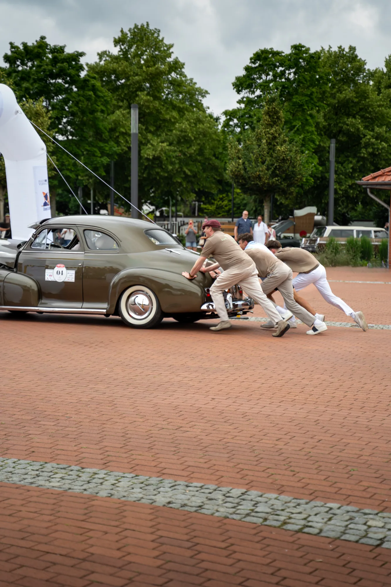 Mehrere Teilnehmer schieben einen Oldtimer auf einem gepflasterten Platz bei den BOC 2025.