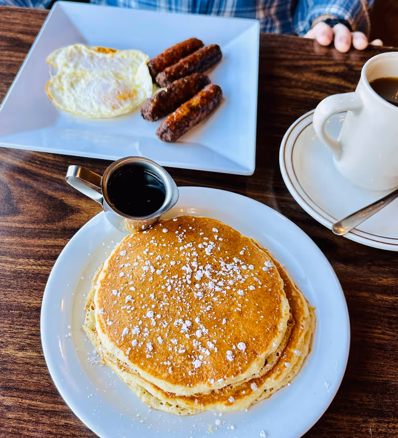 A plate of pancakes with syrup and sausage on a wooden table.