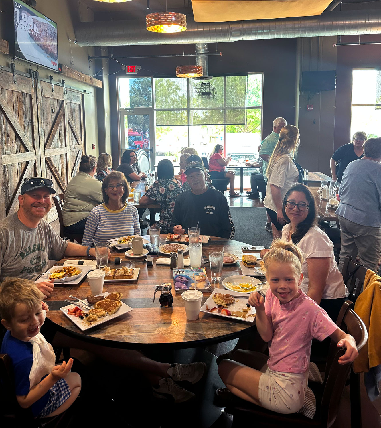 A family of six smiles while enjoying breakfast at a restaurant, seated around a rustic wooden table with plates of pancakes and drinks, creating a warm, cheerful atmosphere.