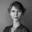 Portrait of a woman with glasses and wavy hair pulled back, wearing a dark blue blouse against a gray background.