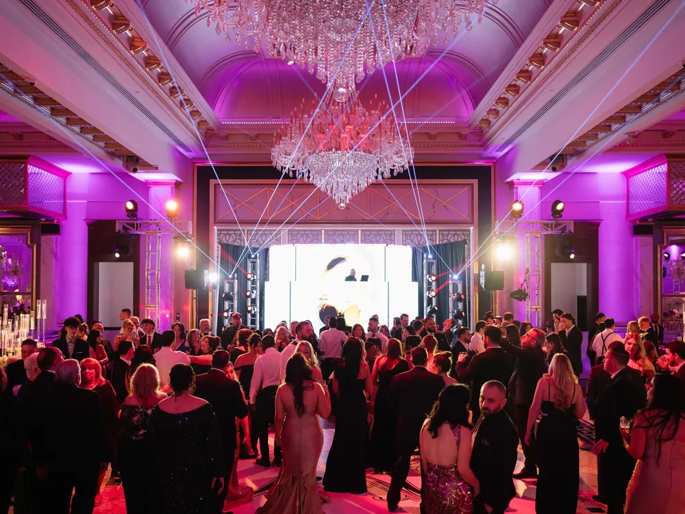 Laser lighting beams sweep across a packed dance floor under crystal chandeliers in a pink-lit ballroom