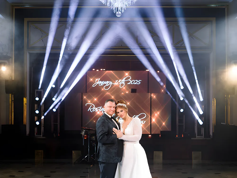 Bride and groom posing under white theatrical lighting beams with a lit LED screen showing their wedding date