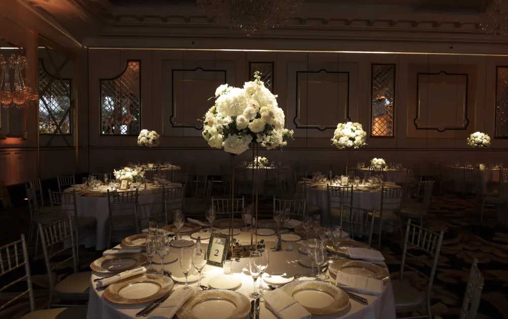 Pin spot lighting highlights white flower centerpieces on round tables in a dimly lit reception room
